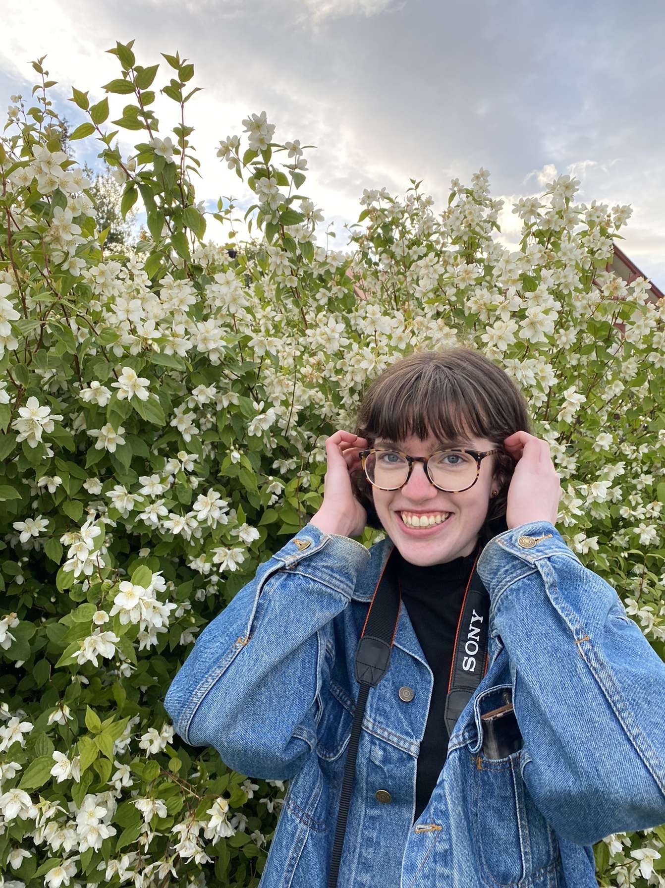 Lilja stands in front of plants with a camera around their neck.